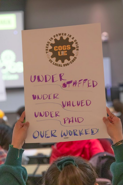 A grad holds a sign reading "Under staffed, under valued, under paid, over worked."